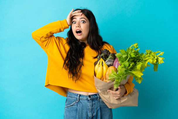 Mujer joven sosteniendo una bolsa de la compra de comestibles aislado sobre fondo azul haciendo gesto sorpresa mientras mira a un lado - Foto, Imagen