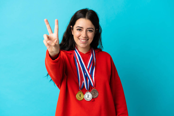 jonge vrouw met medailles geïsoleerd op witte achtergrond glimlachen en tonen overwinning teken - Foto, afbeelding