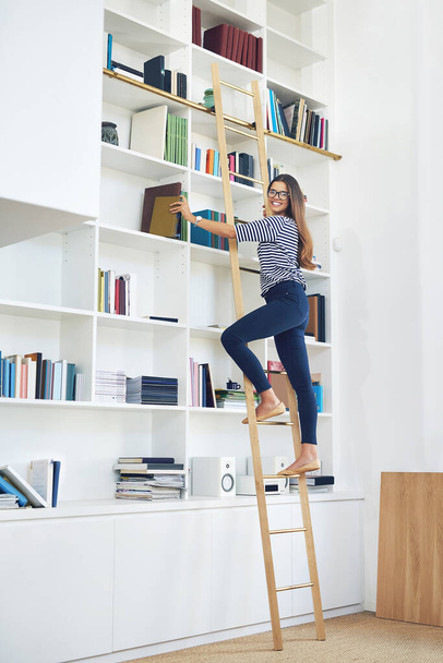 Portrait of a smiling young woman climbing a ladder on a bookshelves at home - Fotó, kép