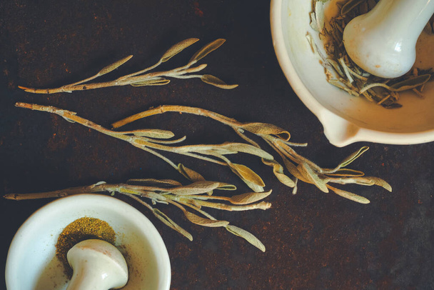 Sementes de cânhamo triturado e ervas de sálvia secas em uma superfície de mesa áspera com uma argamassa de cerâmica branca velha e pilão, vista superior - Foto, Imagem