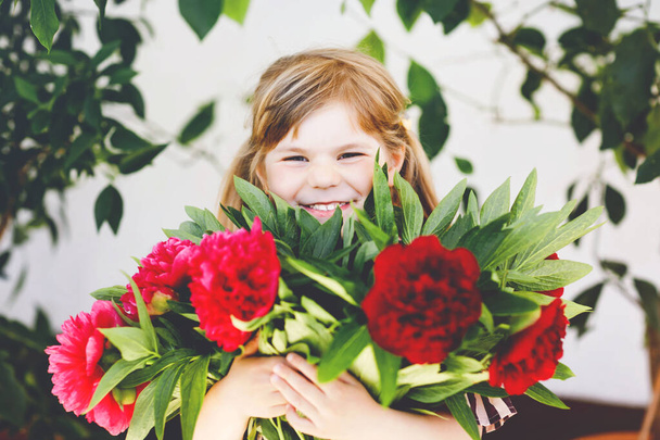 Cute adorable little toddler girl with huge bouquet of blossoming red and pink peony flowers. Portrait of smiling preschool child in domestic garden on warm spring or summer day. Summertime. - Photo, Image
