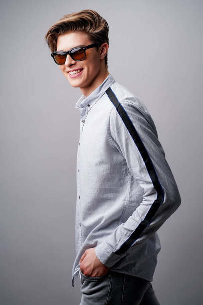 Portrait of smiling handsome guy standing in sunglasses holds arms in pockets, isolated on a gray background - Foto, immagini