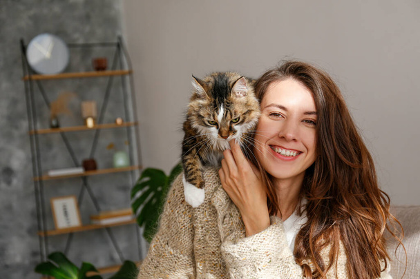 Portrait of young woman in the living room at home with a cute siberian cat. Female hugging her long hair kitty. Background, copy space, close up. Adorable domestic pet concept. - Foto, imagen