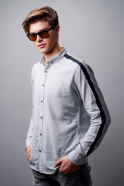 Portrait of handsome guy standing in sunglasses holds arms in pockets, isolated on a gray background - Foto, immagini