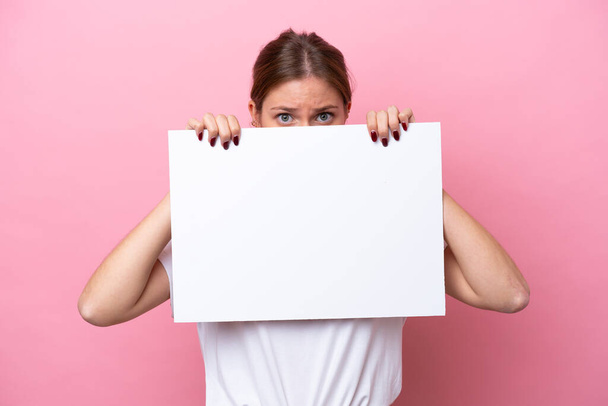 Young caucasian woman isolated on pink background holding an empty placard and hiding behind it - Foto, imagen