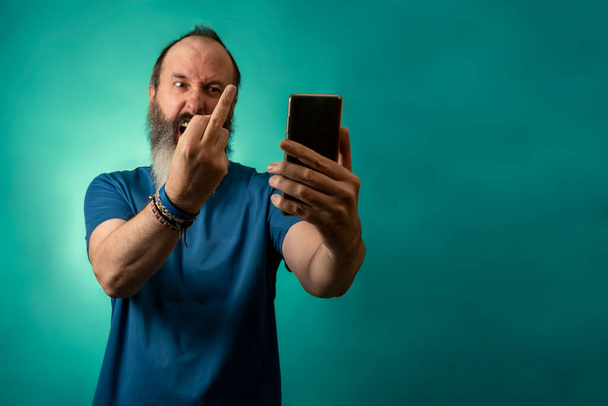 Vousatý muž v neformálním modrém tričku naštvaný, křičící a hrubě ukazující prostředníček do smartphonu - Fotografie, Obrázek