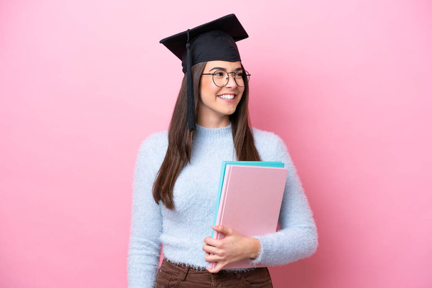 Giovane studentessa brasiliana indossa cappello laureato isolato su sfondo rosa cercando lato - Foto, immagini