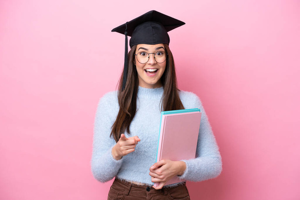 Joven estudiante brasileña con sombrero graduado aislado sobre fondo rosa sorprendida y apuntando hacia delante - Foto, imagen