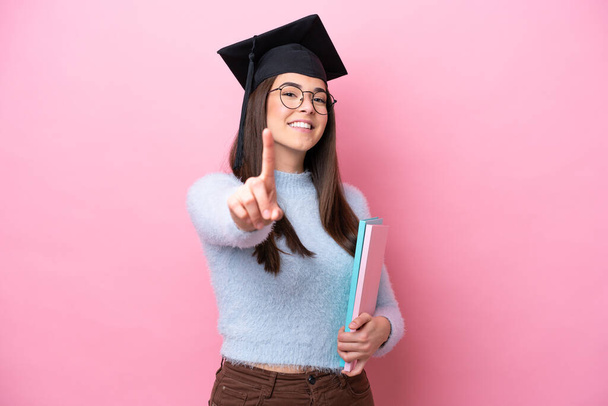 Joven estudiante brasileña con sombrero graduado aislado sobre fondo rosa mostrando y levantando un dedo - Foto, Imagen