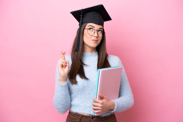 Jeune étudiante brésilienne portant un chapeau gradué isolé sur fond rose avec les doigts croisés et souhaitant le meilleur - Photo, image