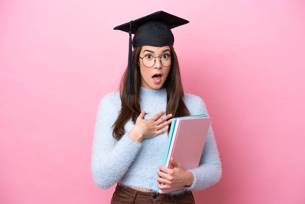 Joven estudiante brasileña con sombrero graduado aislado sobre fondo rosa sorprendida y sorprendida mientras mira a la derecha - Foto, imagen