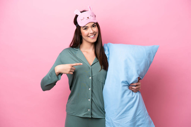 Jovem brasileira isolada sobre fundo rosa em pijama e apontando um travesseiro - Foto, Imagem