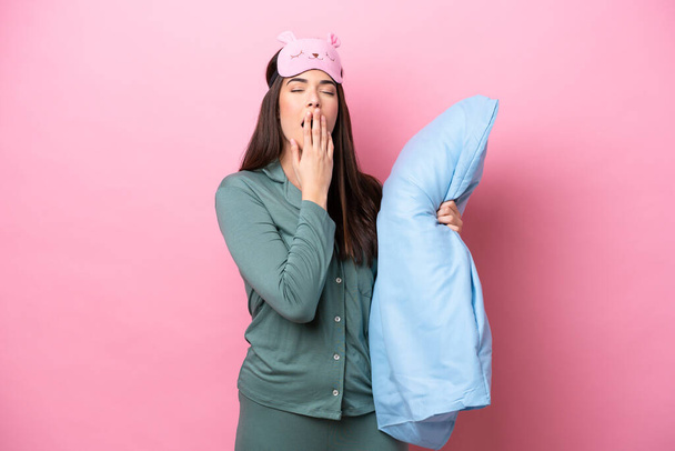 Young Brazilian woman isolated on pink background in pajamas and holding a pillow and yawning - Photo, Image