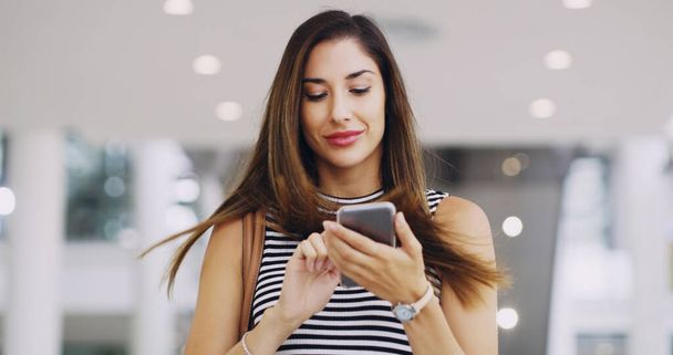 It pays to stay connected. Cropped shot of a young businesswoman using a smartphone while walking through a modern office. - Foto, imagen