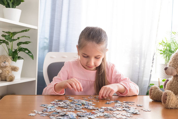 6-7 yaşlarındaki pembe tişörtlü küçük kız evde oyuncak ayıyla bir masada oturuyor ve bulmacaları topluyor.. - Fotoğraf, Görsel