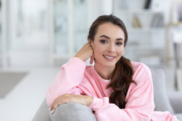 Close up portrait of beautiful calm happy woman sitting on sofa cute smiling. Female student posing indoors in a pink sweatshirt. - Фото, зображення