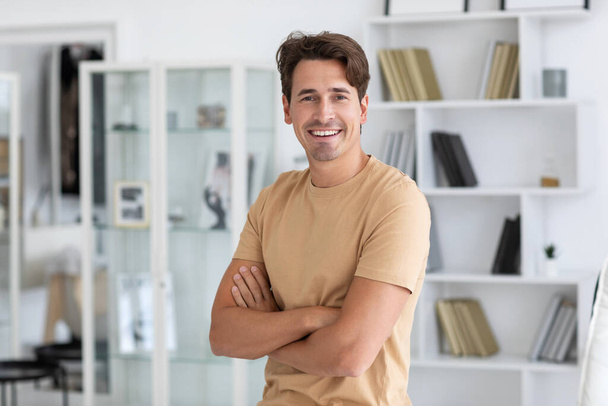 Πορτρέτο ενός όμορφου άντρα σε ένα εσωτερικό γραφείο στο σπίτι, χαμογελαστός τύπος σε μπεζ μπλουζάκι κοιτάζει κάμερα. - Φωτογραφία, εικόνα