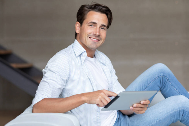 Handsome smiling man with digital tablet on the sofa at home, looking at the camera online shopping and netsurfing. - Photo, Image