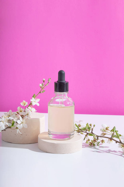 Glass dropper bottle with cosmetic oil on podium with blooming twigs on pink background. Vertical stock photo - Foto, Imagem