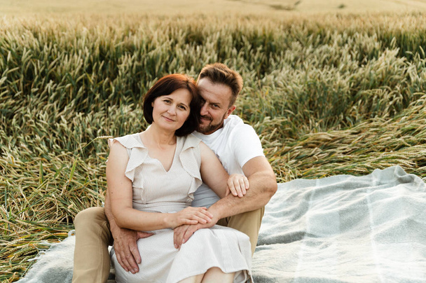 beautiful middle-aged couple in a wheat field sitting on the bedspread and hugging tenderly at sunset. older people in love. lovers having a picnic in the field - Fotoğraf, Görsel