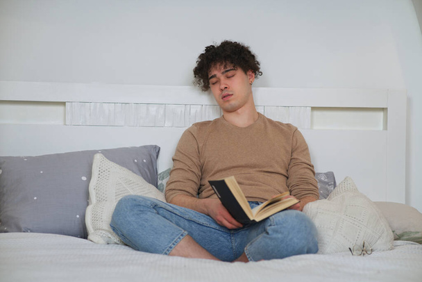 Portrait of handsome tired exhausted sleepy guy, young man is sleeping sitting on cozy bed in bedroom with open book in hands. Reader fall asleep, daydream at home. Student preparing to exam - Photo, Image