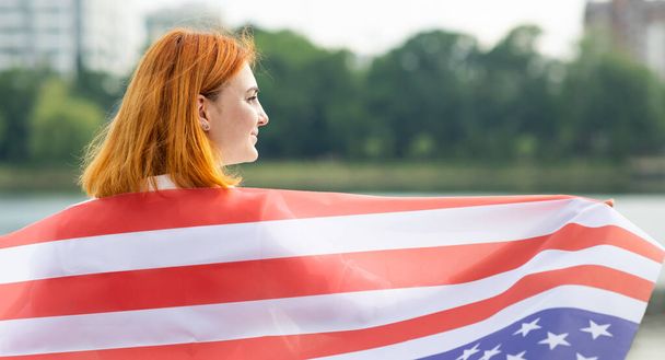 Widok z tyłu szczęśliwej młodej kobiety z flagą USA na ramionach. Pozytywna dziewczyna świętuje dzień niepodległości Stanów Zjednoczonych - Zdjęcie, obraz