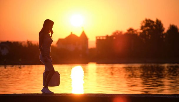 Młoda kobieta spacerująca samotnie po brzegu jeziora w ciepły wieczór. Samotność i relaks w koncepcji przyrody - Zdjęcie, obraz