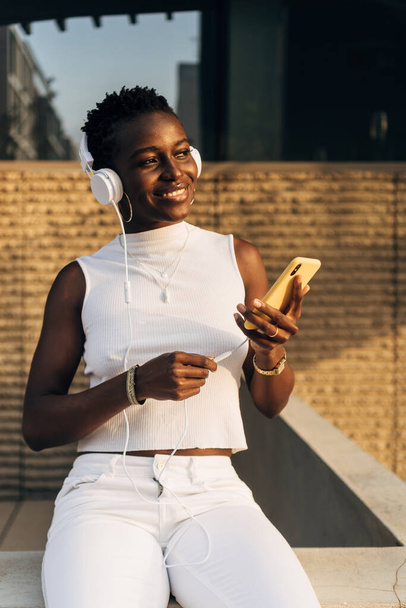 Femme afro-américaine aime écouter de la musique avec un téléphone portable et écouteurs tout en étant assis à l'extérieur. - Photo, image