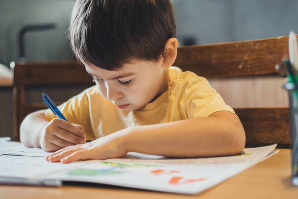 Маленький белый мальчик сидит за столом и делает домашнее задание для начальной школы. Школьный класс. - Фото, изображение