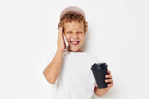Little boy in a white t-shirt cap with a phone in a glass with a drink light background unaltered - Фото, изображение