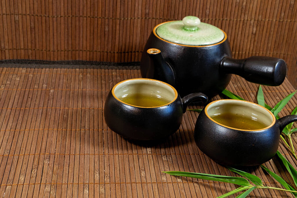 Théière japonaise et tasses à thé vert sur un tapis de bambou avec des feuilles de bambou, - Photo, image