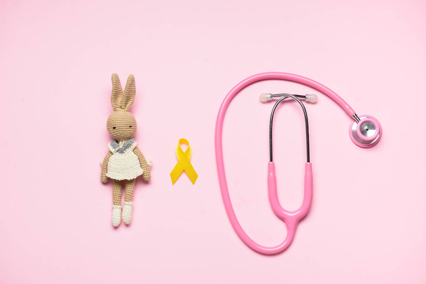 Golden awareness ribbon, toy rabbit and stethoscope on color background. International Childhood Cancer Day - Photo, Image