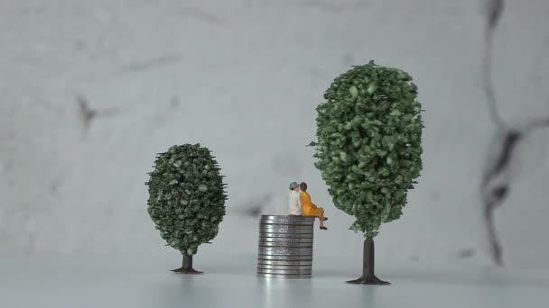Ouderdom miniatuur mensen op een stapel munten tussen miniatuur bomen. Het concept van een vergrijzende samenleving en de stijgende sociale kosten. - Video