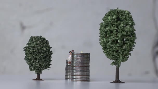 A miniature man and a miniature woman sitting with a baby on top of a stack of coins. The concept of miniature people and social problems. - Footage, Video