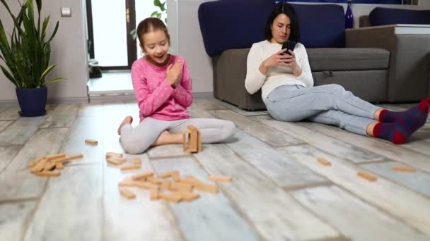 Indifferent mother use mobile phone, daughter play wooden construction on floor at home, busy woman is working at home little daughter nearby, Dont have enough time for child, phone addict parents - 映像、動画