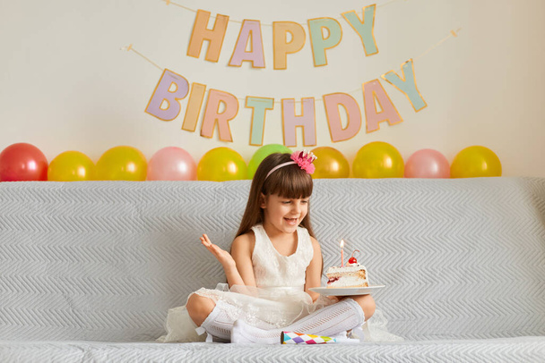 Horizontal shot of happy smiling little girl wearing white dress sitting on sofa with birthday cake and burning candle, rejoicing her holiday, raised arm, expressing happiness. - Valokuva, kuva