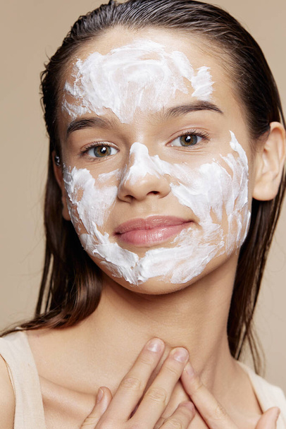 sorridente giovane donna cura della pelle utilizzando maschera bianca sul viso sfondo beige - Foto, immagini