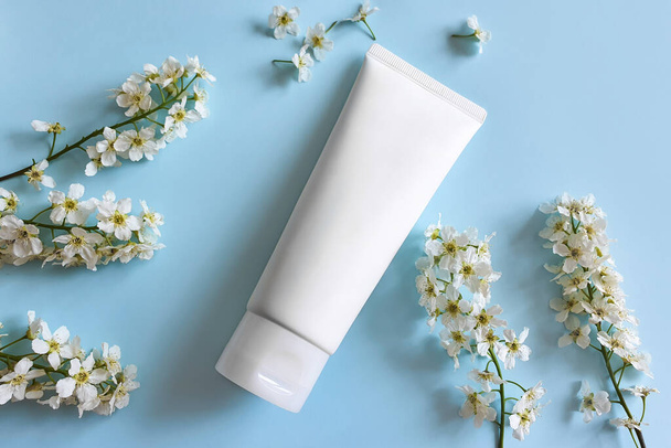 White squeeze bottle plastic tube for branding of medicine or cosmetics - cream, gel, skincare, moisturize. Cosmetic container and white bird cherry flowers on blue background. Top view, mockup. - Photo, Image