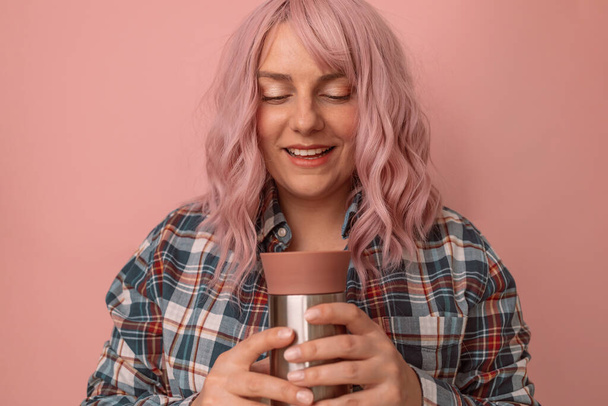 Affascinante giovane donna caucasica con i capelli rosa che tiene un thermos con una bevanda calda su sfondo rosa pastello - Foto, immagini