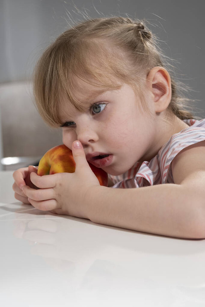 little girl with a big red apple is sitting at the table in the room - Photo, Image