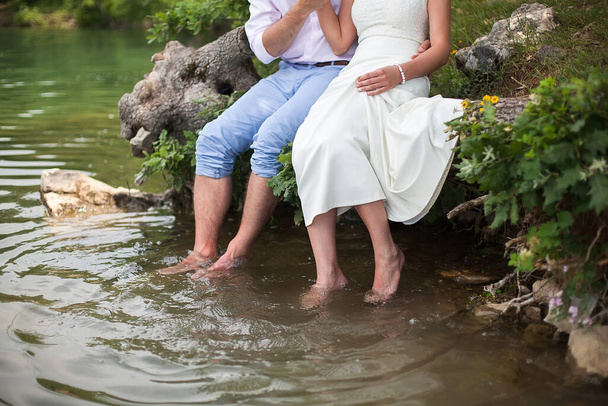 The bride and groom sitting on the shore of the lake. Foot splashing in the water - Fotoğraf, Görsel