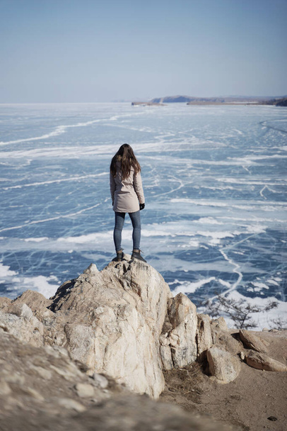 Winter Lake Baikal. Woman Standing on a Cliff and Looking on a Frozen Baikal lake. Olkhon Island, Russia, Siberia - Foto, afbeelding