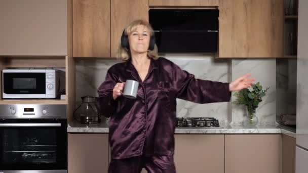 Mujer rubia madura baila en auriculares sosteniendo una taza de té - Metraje, vídeo