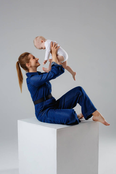 Cute baby in his mother's arms on a gray background in the studio. A mother in denim clothes lovingly embraces a child. The concept of motherhood. Unconditional love. - Photo, image