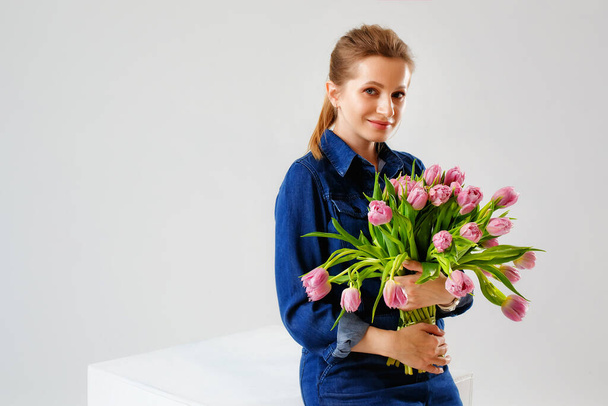 Portré egy fiatal nőről farmer kezeslábasban, egy nagy csokor rózsaszín tulipánnal, szürke háttérrel a stúdióban. Szőke virágokkal, mint ajándék a nők napjára. - Fotó, kép