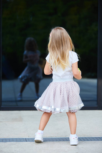 白いTシャツとピンクのスカートの小さなブロンドの女の子は、ガラスの表面に彼女の反射を見ています。子供時代と大人の模倣の概念. - 写真・画像