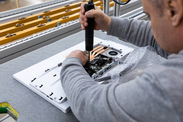 灰色のセーターを着た成人男性の写真で、モノブロックの組立工場のコンベアベルトにコンピュータモニタシステムブロックを組み立てます。エンジニアは現代のハイテク工場で働いています - 写真・画像