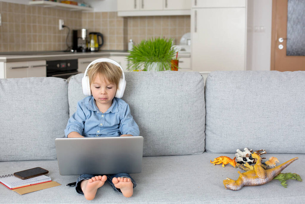 Sweet blond child, boy in casual clothes, woring on computed at home, with headphones and phone - Fotó, kép