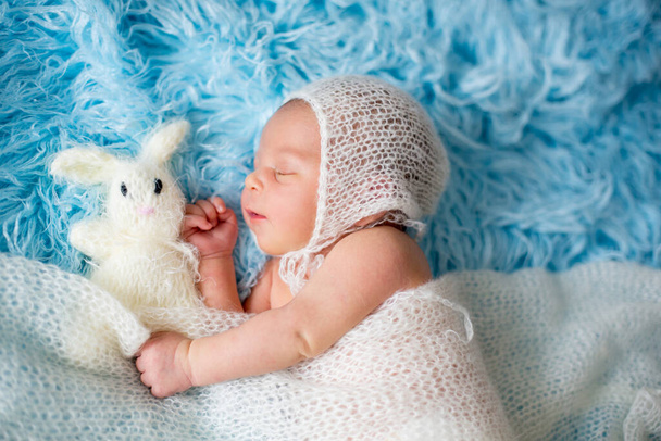 Little cute newborn baby boy, sleeping wrapped in white wrap, holding little knitted toy - Fotoğraf, Görsel