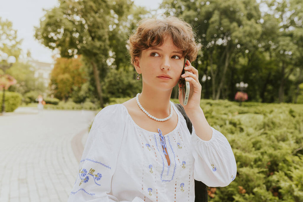 νεαρή κοπέλα φοιτητής ή μαθήτρια με καστανά σγουρά μαλλιά, σε κεντημένο εθνικό εθνικό λευκό πουκάμισο, καλεί τηλέφωνο με επιφυλακτική έκφραση, ενώ το περπάτημα στο πάρκο την άνοιξη ηλιόλουστη μέρα. Συναγερμός νεαρή γυναίκα διεσπαρμένο πάρκο - Φωτογραφία, εικόνα